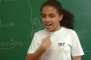 Picture of a girl from Brazil standing in front of a blackboard using sign language.