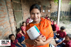 Ruma stands confidently holding a text book with Bangla characters. She is wearing a beautiful orange beaded and embroidered shawl and dress. Behind her, a group of around 10 young Bangladeshi boys and girls in school uniforms sit on the ground outside, next to a brick building, ready for a lesson, together with some young women and young children.