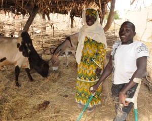 Picture of a man in Niger, who uses crutches, and a woman, standing next to a goat and a donkey. The animals are under a thatched roof shelter.