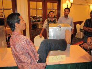 A man from Indonesia who has no arms, uses his foot to place a ballot paper in the locked metal ballot box. The ballot box is high on a table. Three men are watching from the other side of the table. 
