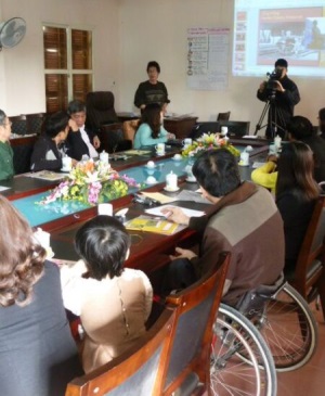 A number of men and women from Vietnam, including a wheelchair user (third from the left) sit around a table participating in discussion, and watching a powerpoint presentation.