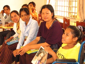 Picture of a group of 7 women from Cambodia sitting in a meeting room.  The woman to the front of the picture is a wheelchair user. She is holding a picture and speaking. The other women are all listening to her. 