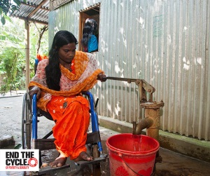 Picture of a woman in Bangladesh who is a wheelchair user at an accessible water pump. The water pump is low enough to be reached sitting down, and has a long handle which she is using to collect water in a plastic bucket underneath. 
